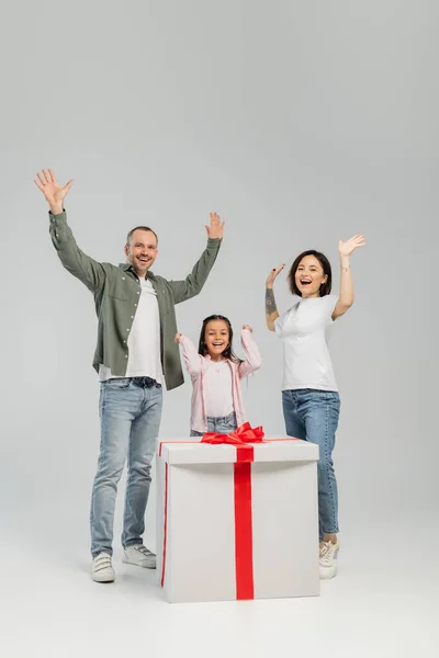Excited parents in casual clothes waving hands at camera near preteen daughter and big present during international child protection day celebration on grey background — Stock Photo