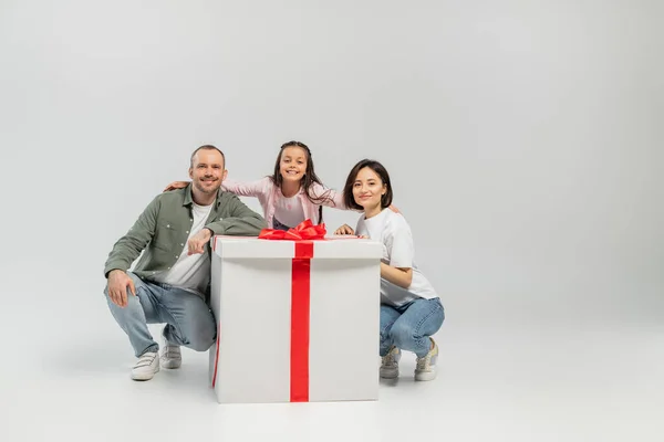 Positives Mädchen umarmt Eltern in lässiger Kleidung und blickt in die Kamera in der Nähe einer großen Geschenkbox während der internationalen Kindertagsfeier auf grauem Hintergrund — Stockfoto