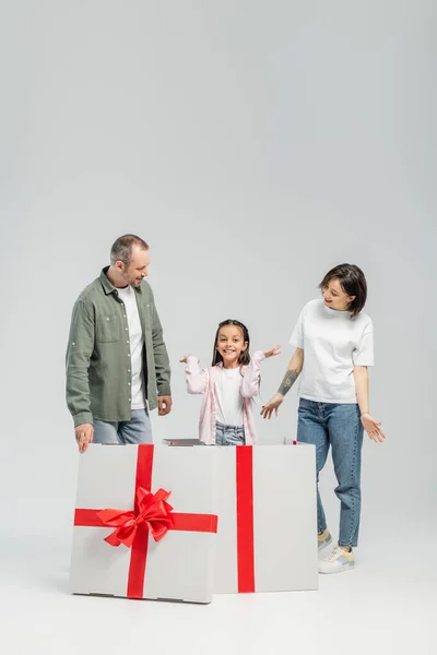 Full length of cheerful parents looking at preteen daughter looking at camera while standing inside of big present box during child protection day on grey background — Stock Photo