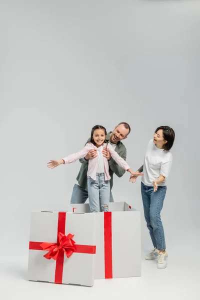 Веселый мужчина вынимает дочку большой подарок коробка с красной лентой рядом счастливая жена с татуировкой на руке во время празднования Дня защиты детей на сером фоне — стоковое фото
