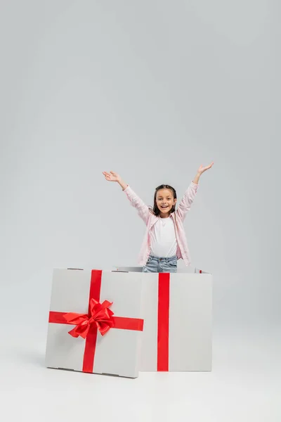 Aufgeregtes und fröhliches Mädchen in lässiger Kleidung, das in die Kamera blickt, während es in einer großen Geschenkbox während des internationalen Kindertages auf grauem Hintergrund steht — Stockfoto