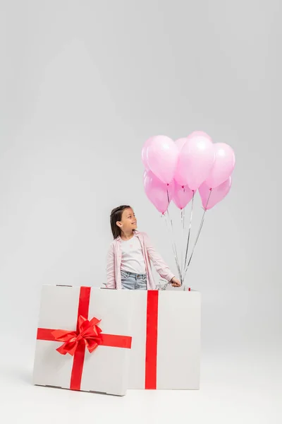Niño preadolescente sonriente con ropa casual mirando globos rosados mientras está de pie en una gran caja de regalo durante la celebración del día de los niños felices sobre un fondo gris - foto de stock