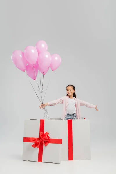 Niño preadolescente despreocupado con ropa casual mirando a la cámara mientras sostiene globos festivos rosados mientras está de pie en un gran regalo durante la celebración del día internacional de los niños sobre un fondo gris - foto de stock
