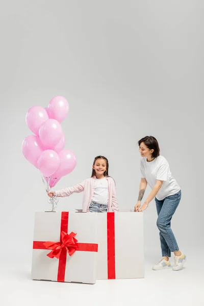 Улыбающаяся татуированная женщина в повседневной одежде смотрит на дочь с воздушными шарами и большой коробкой подарков во время празднования Дня детей на сером фоне — стоковое фото