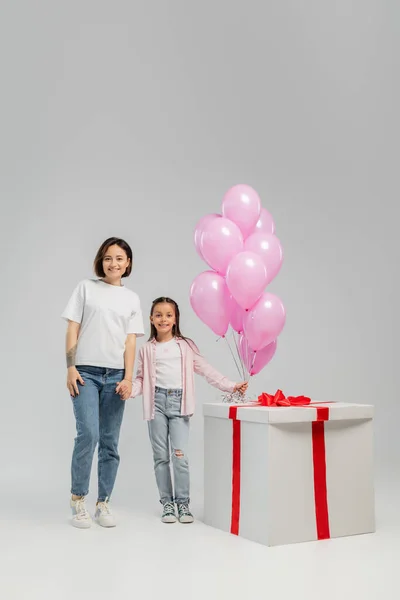 Comprimento total de mãe tatuada sorridente segurando a mão da filha pré-adolescente perto de balões rosa e grande caixa de presente durante a celebração do dia das crianças no fundo cinza — Fotografia de Stock