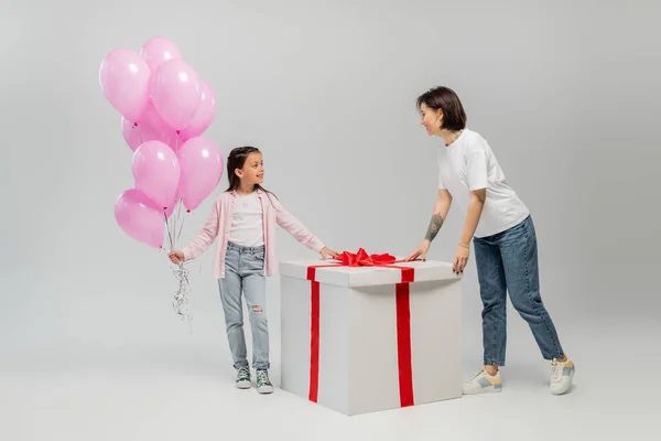 Longitud completa de niña preadolescente sonriente sosteniendo globos rosados y mirando a la mamá tatuada cerca de la caja de regalo grande durante la celebración del día feliz de los niños sobre fondo gris - foto de stock