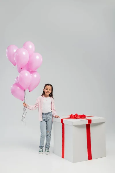 Full length of carefree preteen kid in casual clothes looking at camera while holding pink balloons near big present during child protection day on grey background — Stock Photo