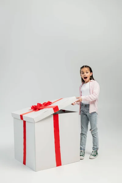 Full length of shocked preteen girl in casual clothes looking at camera while opening big gift box during child protection day celebration on grey background — Stock Photo
