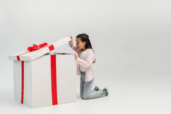 Side view of shocked preteen kid in casual clothes opening and looking at big present box during international children day celebration on grey background — Stock Photo