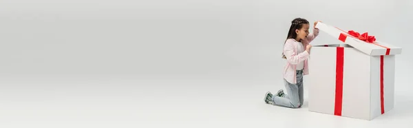 Vue latérale de la préadolescente choquée en vêtements décontractés ouvrant une grande boîte-cadeau avec arc lors de la célébration internationale de la journée de protection des enfants sur fond gris, bannière — Photo de stock