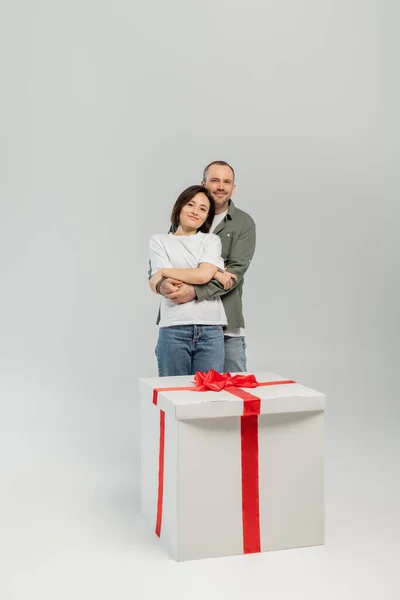 Smiling man in casual clothes hugging wife and looking at camera near big present with bow during international children day celebration on grey background — Stock Photo