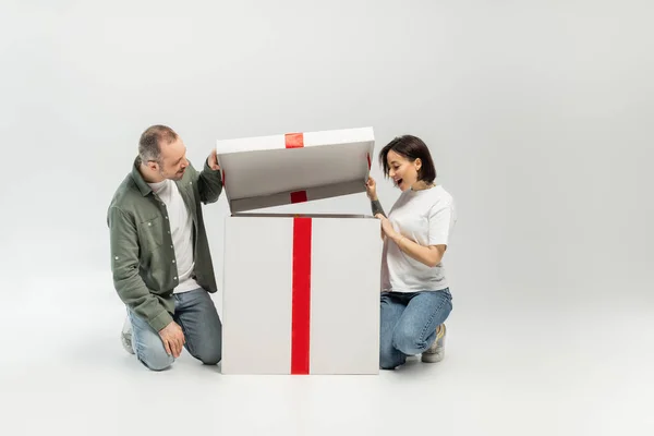 Возбужденная татуированная женщина смотрит на открытую большую коробку подарков рядом с мужем во время празднования Дня защиты детей на сером фоне с копировальным пространством — стоковое фото