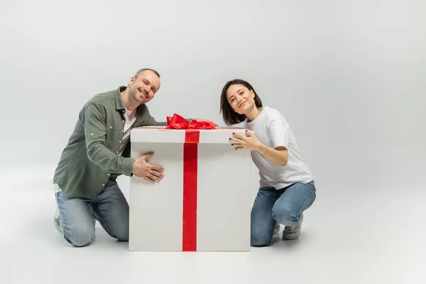 Улыбающаяся взрослая пара в повседневной одежде смотрит в камеру, обнимая большую коробку подарков и глядя на камеру во время дня защиты детей на сером фоне — стоковое фото