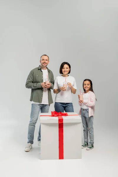 Positive Familie mit frühgeborener Tochter, die Milchshakes in die Kamera hält und in der Nähe einer großen Geschenkbox während der Kinderschutztagsfeier auf grauem Hintergrund blickt — Stockfoto