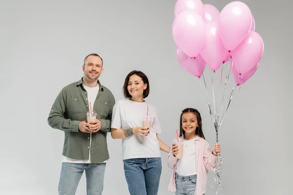 Los padres sonrientes y la hija preadolescente sosteniendo globos rosados y batidos mientras miran a la cámara durante la celebración del día internacional de los niños aislados en gris - foto de stock