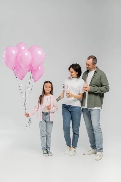 Улыбающиеся родители, держащие молочные коктейли и смотрящие на дочь-подростка с воздушными шарами во время празднования Международного дня защиты детей на сером фоне — стоковое фото