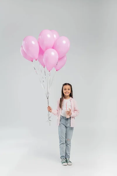 Voller Länge lächelnde Preteen Mädchen in lässiger Kleidung mit rosa Luftballons und Milchshake, während sie während der Kinderschutztag-Feier auf grauem Hintergrund in die Kamera schauen — Stockfoto