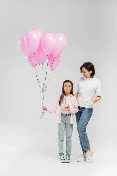 Full length of smiling mother in casual clothes holding milkshake and hugging preteen daughter with pink balloons during child protection day on grey background — Stock Photo