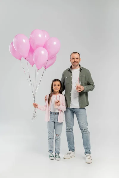 Padre sonriente e hija preadolescente con ropa casual sosteniendo batidos y globos rosados mientras celebra el día de la protección del niño y de pie sobre un fondo gris - foto de stock