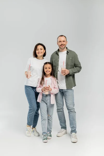 Full length of smiling family with preteen daughter in casual clothes holding milkshakes in plastic cups and looking at camera during children day celebration on grey background — Stock Photo