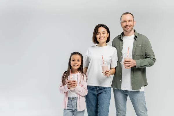 Parents positifs en vêtements décontractés regardant la caméra près de la fille avec milkshake dans une tasse en plastique lors de la célébration de la journée internationale des enfants isolé sur gris avec espace de copie — Photo de stock