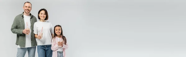 Улыбающиеся взрослые родители в повседневной одежде, держащие молочные коктейли рядом с ребенком-подростком и смотрящие в камеру в международный день защиты детей, изолированные на сером с копировальным пространством, баннер — стоковое фото