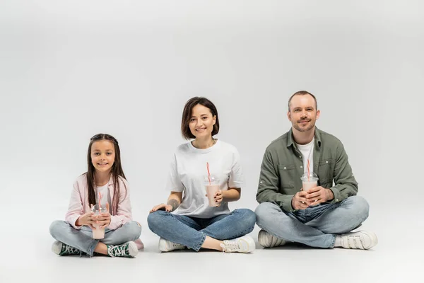 Smiling adult parents in casual clothes looking at camera while holding plastic cups with milkshakes near preteen daughter during international children day celebration and sitting on grey background — Stock Photo
