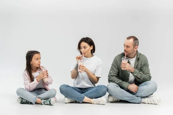 Дорослі батьки в повсякденному одязі п'ють молочні коктейлі з пластикових чашок з п'яними соломинками і дивляться на доньку під час святкування Дня захисту дітей, сидячи на сірому фоні — стокове фото