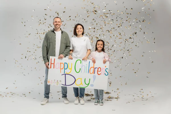 Full length of cheerful family with preteen daughter in casual clothes holding placard with happy children's day lettering and standing under falling festive confetti on grey background — Stock Photo