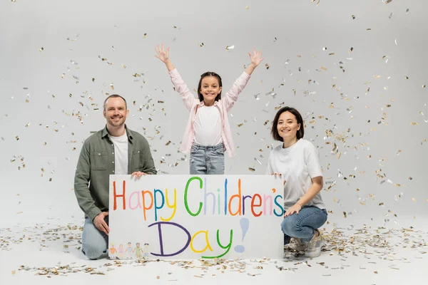 Excited preteen girl in casual clothes waving hands at camera while standing near parent holding placard with happy children's day lettering and under falling festive confetti on grey background — Stock Photo
