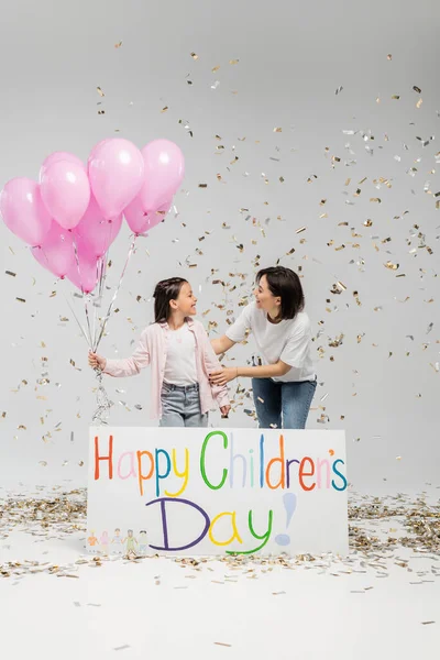 Side view of cheerful woman in casual clothes hugging preteen daughter with pink balloons while standing together near placard with happy children day under falling confetti on grey background — Stock Photo