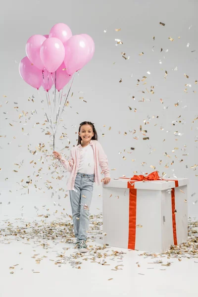 Full length of carefree preteen girl in casual clothes holding pink balloons and looking at camera near big gift box while standing under confetti during child protection day celebration on grey background — Stock Photo