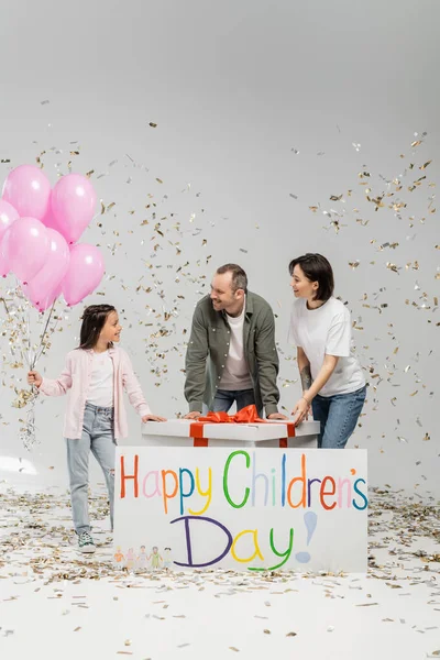 Positive parents looking together at preteen daughter with pink balloons near big present, placard with happy children's day lettering and falling confetti during celebration on grey background — Stock Photo