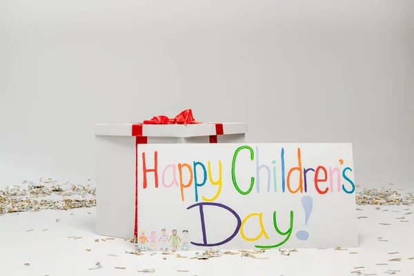 Placard with colorful happy children's day lettering near big gift box with bow on grey background with festive confetti — Stock Photo