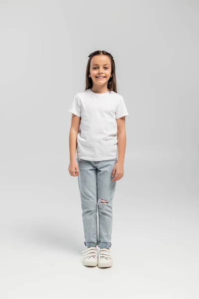 Полная длина веселой и юной девушки в белой футболке и джинсах, смотрящей в камеру во время празднования глобального дня защиты детей и стоящей на сером фоне — стоковое фото