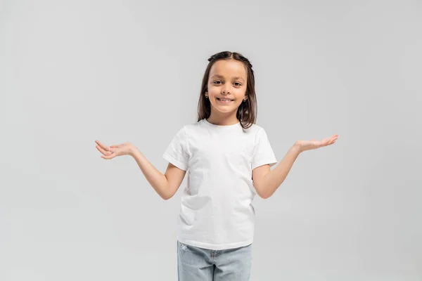 Sonriente niña preadolescente morena en camiseta blanca y jeans mirando a la cámara y mostrando un gesto de encogimiento mientras celebra el día de la protección infantil y de pie aislado en gris - foto de stock