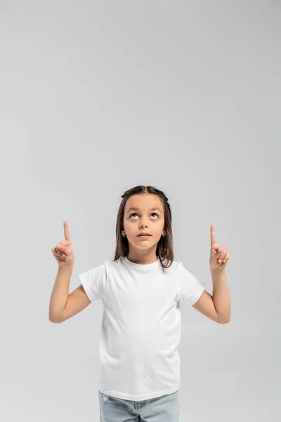 Focused preteen girl in white t-shirt and jeans pointing with fingers and looking up during international child protection day celebration isolated on grey with copy space — Stock Photo