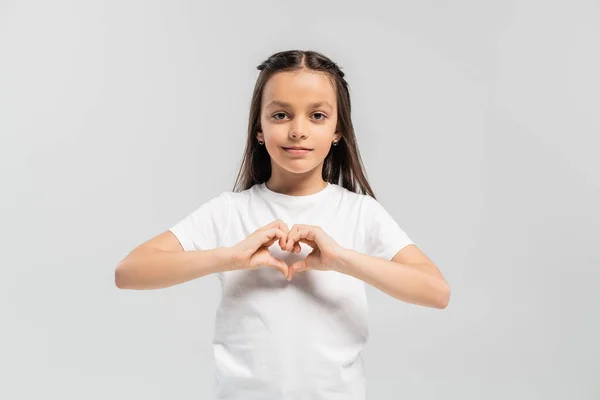 Счастливая девочка-подросток с длинными волосами брюнетки стоя в белой футболке и показывая жест сердца с руками изолированы на сером фоне, День защиты детей праздник — стоковое фото
