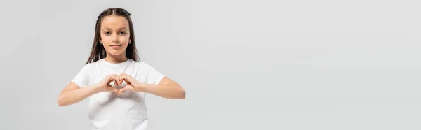 Felice preteen girl con lunghi capelli castani in piedi in t-shirt bianca e mostrando il gesto del cuore con le mani isolate su sfondo grigio, vacanza di protezione del bambino, banner — Foto stock