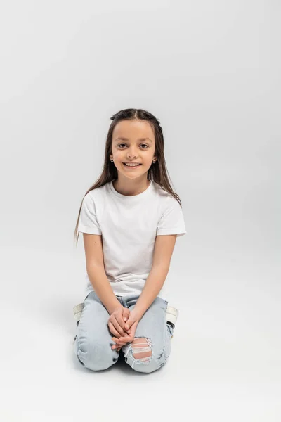 Menina pré-adolescente alegre com cabelos longos morena sentado em t-shirt branca e jeans jeans jeans azul e sorrindo isolado em fundo cinza, feriado dia de proteção infantil — Fotografia de Stock