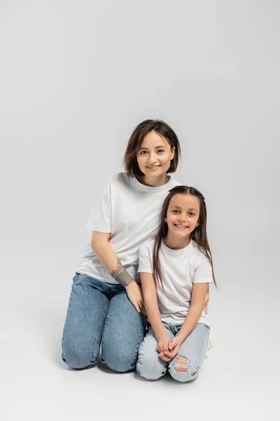 Happy mother with tattoo on hand and short hair hugging preteen brunette daughter while sitting together in white t-shirts and blue denim jeans on grey background, child protection day — Stock Photo