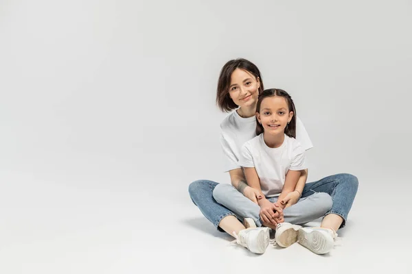 Positive mother with tattoo and short hair hugging preteen brunette daughter while sitting together in white t-shirts and blue denim jeans on grey background, child protection day — Stock Photo