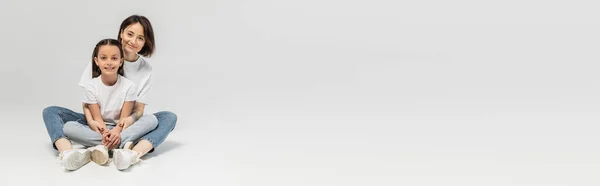 Mãe feliz com tatuagem na mão e cabelo curto abraçando a filha pré-adolescente enquanto sentados juntos em camisetas brancas e jeans jeans jeans azul no fundo cinza, dia de proteção da criança, banner — Fotografia de Stock