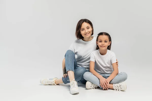 Happy woman with tattoo on hand and short hair hugging preteen daughter while sitting together in white t-shirts and blue denim jeans on grey background, international children's day — Stock Photo