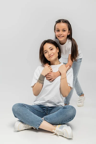 Brunette preteen kid hugging happy mother with tattoo on hand and short hair while sitting together in white t-shirts and blue denim jeans on grey background, international children's day — Stock Photo
