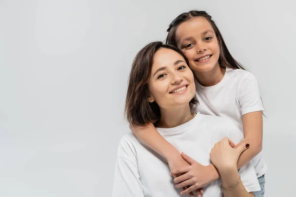 Brunette preteen kid hugging happy mother with short hair while posing together in white t-shirts and looking at camera on grey background, international children's day — Stock Photo