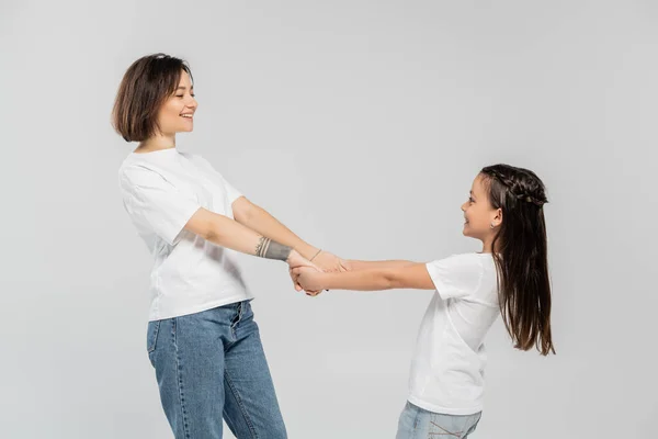 Mãe feliz com tatuagem na mão e cabelo curto de mãos dadas com a filha alegre enquanto estão juntos em camisetas brancas e jeans jeans jeans azul no fundo cinza, dia de proteção da criança — Fotografia de Stock