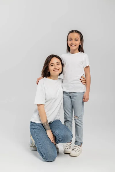 Full length of happy mother with tattoo on hand and short hair hugging preteen daughter while posing together in white t-shirts and blue denim jeans on grey background — Stock Photo