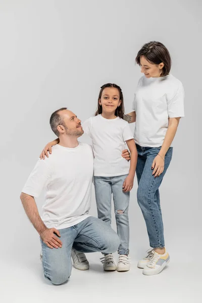 Father and mother with short hair and tattoo on hand looking at cheerful preteen daughter while standing together in white t-shirts and blue denim jeans on grey background, child protection day — Stock Photo