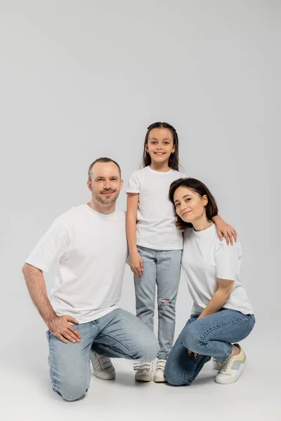 Joyous father and mother with short hair sitting near cheerful preteen daughter while posing together in white t-shirts and blue denim jeans on grey background, Happy children's day — Stock Photo